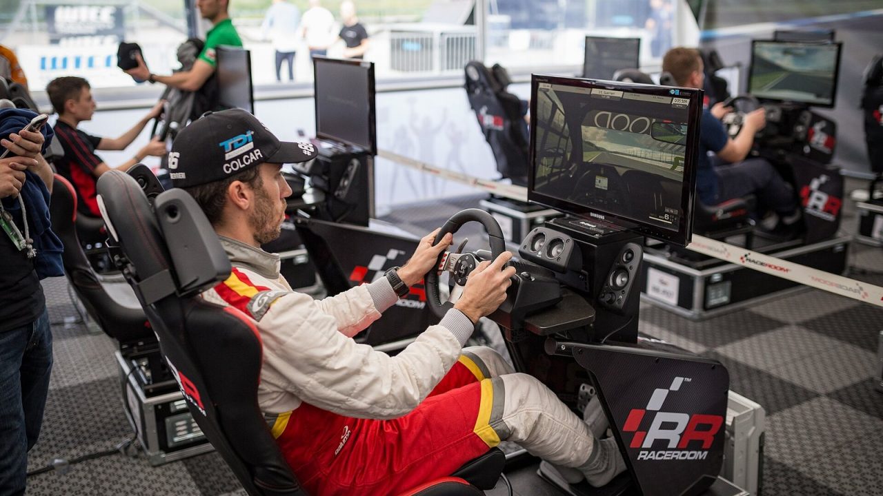 Racing driver in team overalls at a sim racing rig with wheel and screen at an event