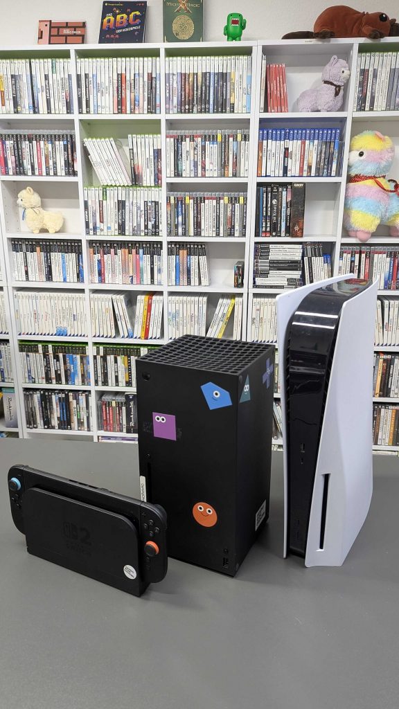 Three current-generation game consoles on a table in front of a shelf of video games